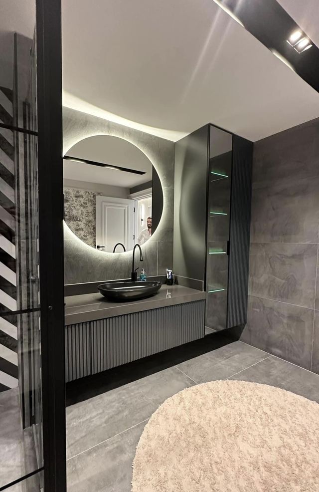 Yuvarlak ışıklı ayna, dikey çıtalı asma lavabo dolabı ve siyah çerçeveli cam raflı dolap