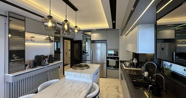 Mermer adalı, sarkıt aydınlatmalı mutfak; arkada vitrinli dolaplar ve gömme cihazlar